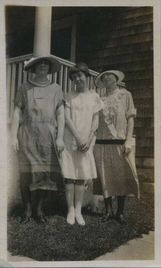 Untitled [Three women stand in front of porch, two on the sides wear hats, woman in center holds her hands in front of her body]