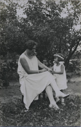 Untitled [Woman sits on chair and holds child's hands]