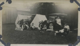 Untitled [Three women, three men, and two children sit on lawn, two cars and two tents in background]