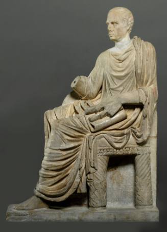 Gravestone in high relief: seated togate male, facing left, his left hand clasping and holding a scroll; another scroll in his right hand