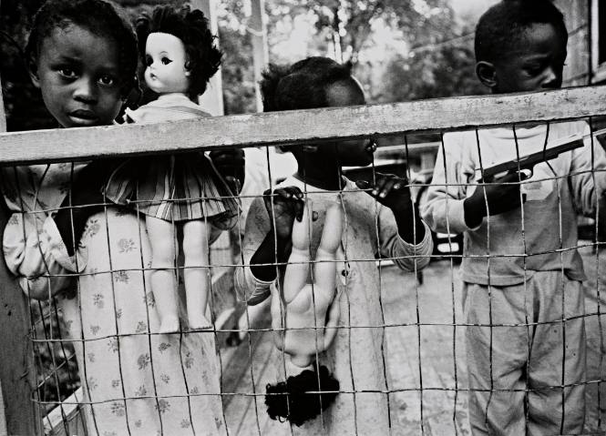 Little girls with dolls, boy with gun, behind wire fence