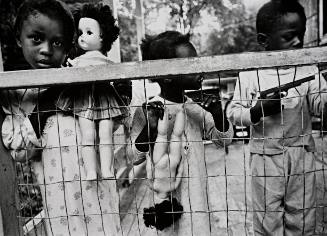 Little girls with dolls, boy with gun, behind wire fence