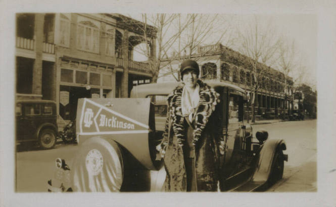Untitled [Woman in fur coat and hat stands in front of car with Dickinson banner]