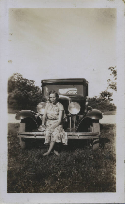 Untitled [Woman in floral dress sits on car's front fender]