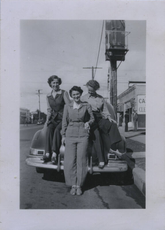 Untitled [Two women in coats and pants sit on a car's back fender, one woman stands on street holding a cigarette]