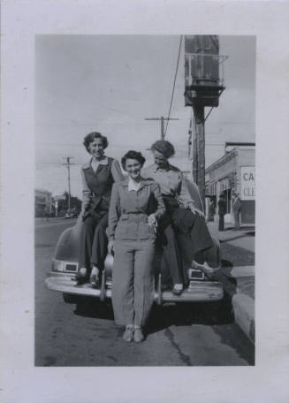 Untitled [Two women in coats and pants sit on a car's back fender, one woman stands on street holding a cigarette]