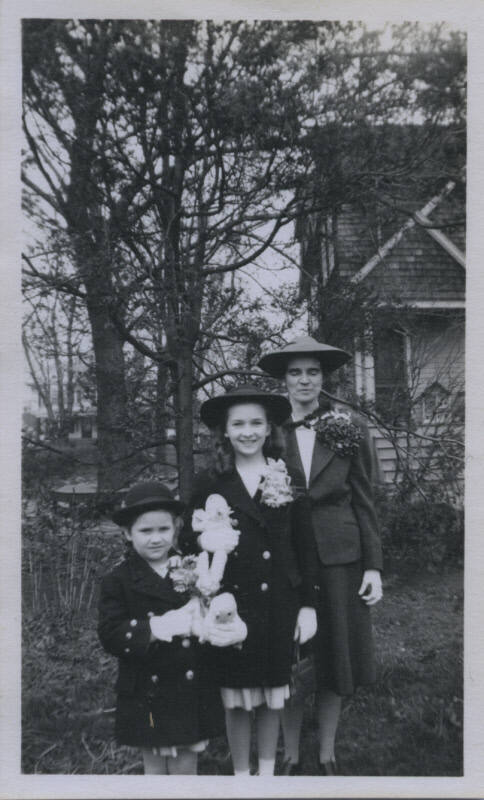 Untitled [Three hatted women stand youngest to oldest, left to right, all wearing flowers on their coats]