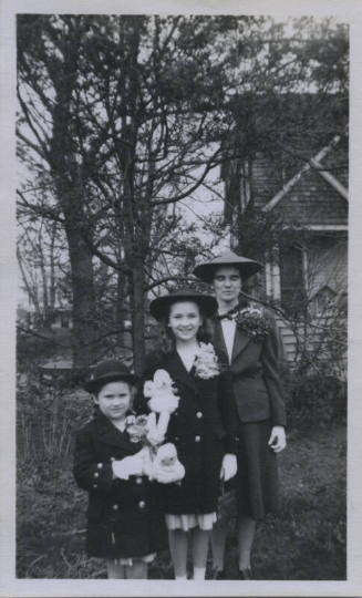 Untitled [Three hatted women stand youngest to oldest, left to right, all wearing flowers on their coats]