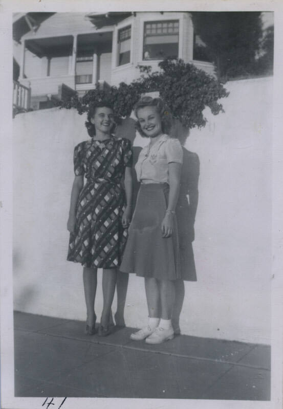 Untitled [Two women stand in front of blank wall, atop which a vine grows]