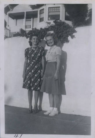 Untitled [Two women stand in front of blank wall, atop which a vine grows]