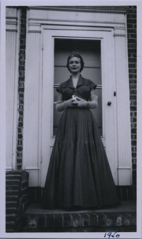Untitled [Woman in long dark dress and gloves stands in front of doorway with hands clasped together]
