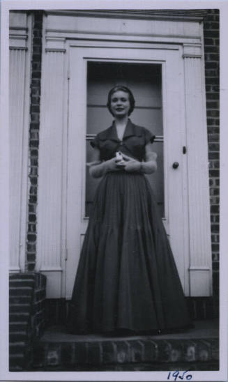 Untitled [Woman in long dark dress and gloves stands in front of doorway with hands clasped together]