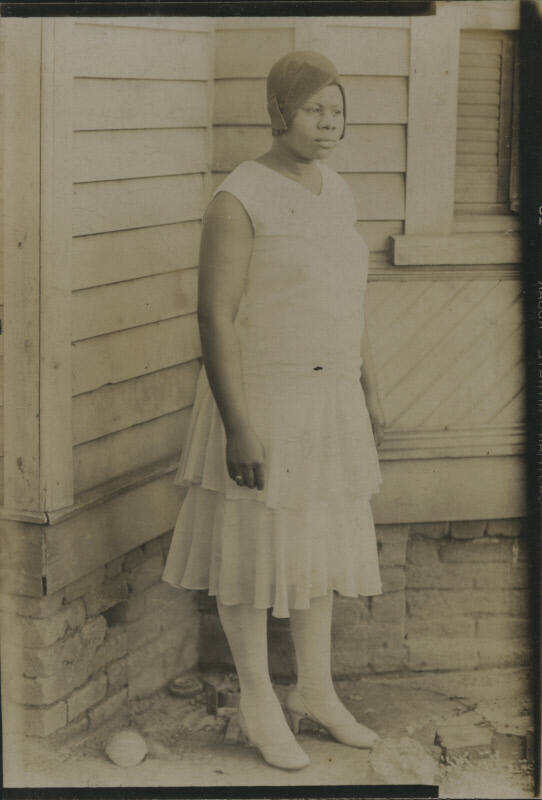 Untitled [Woman in hat and dress looks out, a building behind her]