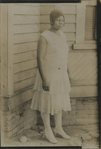 Untitled [Woman in hat and dress looks out, a building behind her]