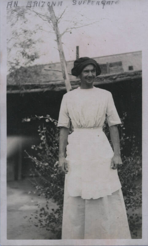 Untitled [Man stands outside house, wears apron and hat]