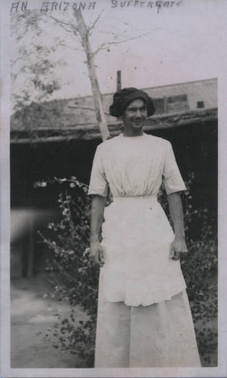 Untitled [Man stands outside house, wears apron and hat]