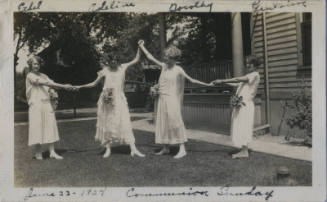 Untitled [Four women in long light gowns punctuated with flower applications hold each other's hands, arms outstretched]