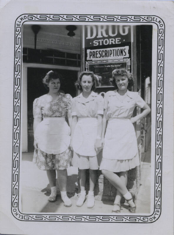 Untitled [Three women wearing aprons stand in front of drug store sign]
