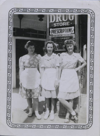 Untitled [Three women wearing aprons stand in front of drug store sign]