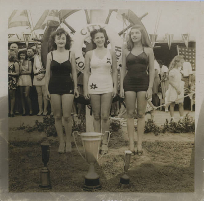 Untitled [Three women stand in front of giant spoked wheel, three trophies in front of them]