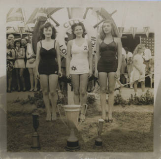 Untitled [Three women stand in front of giant spoked wheel, three trophies in front of them]