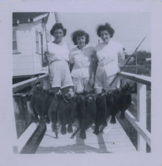 Untitled [Three women on a dock hold up a line of fish]