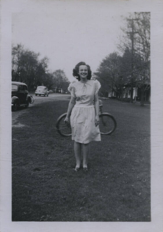 Untitled [Woman stands in garden, bicycle in the background]