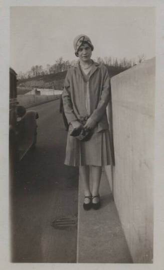Untitled [Woman stands on side of road, holds purse with both hands in front of her]