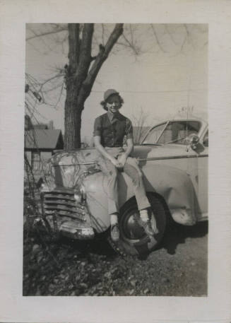 Untitled [Someone sits atop car hood, wears vest and hat]