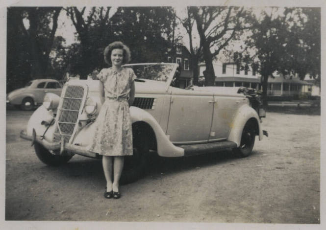 Untitled [Woman in floral dress stands in front of car, arms behind her back]