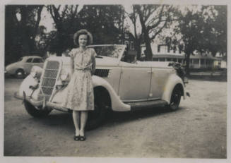 Untitled [Woman in floral dress stands in front of car, arms behind her back]