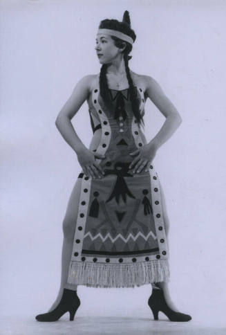 Untitled [Woman wears a cross necklace, feather headdress and a patterned tunic, props hands on hips]