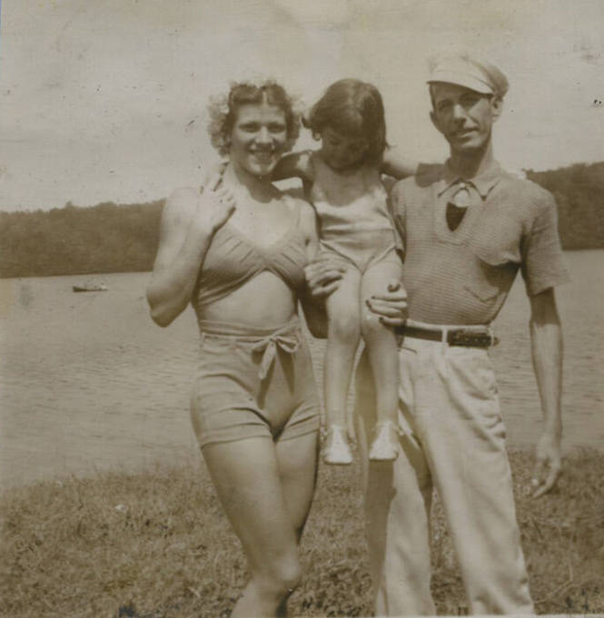 Untitled [Woman, child, and man stand in front of body of water, woman and man hold up child by their legs]
