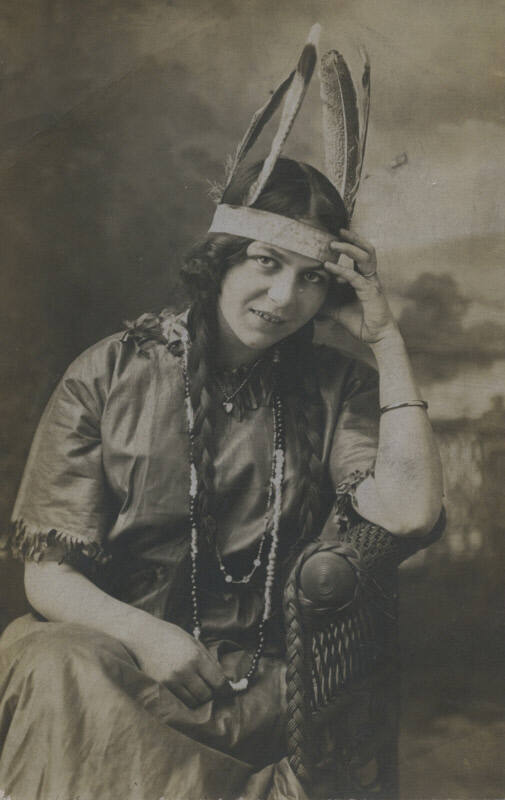 Untitled [Woman sits on wicker chair, wears heart necklace, wedding band, and feathered headdress, rests head on hand]