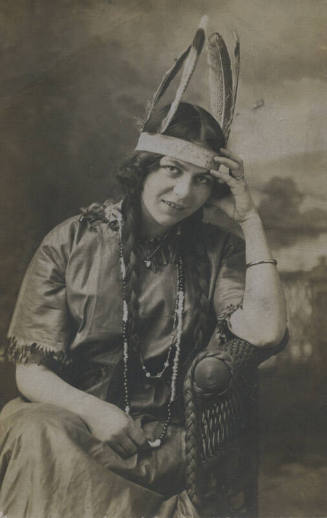Untitled [Woman sits on wicker chair, wears heart necklace, wedding band, and feathered headdress, rests head on hand]