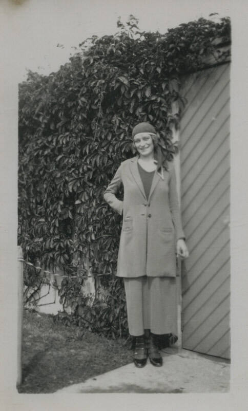 Untitled [Woman stands in front of gate, wall of leaves in the background]