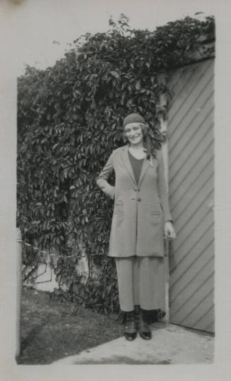 Untitled [Woman stands in front of gate, wall of leaves in the background]