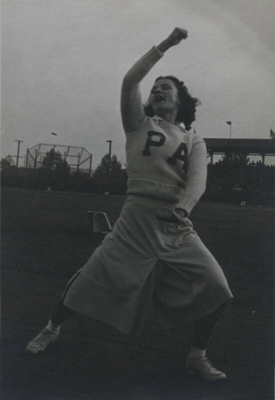 Untitled [Woman holds one arm up and one down in athletic field, wears sweater that reads PA]