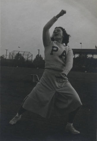 Untitled [Woman holds one arm up and one down in athletic field, wears sweater that reads PA]