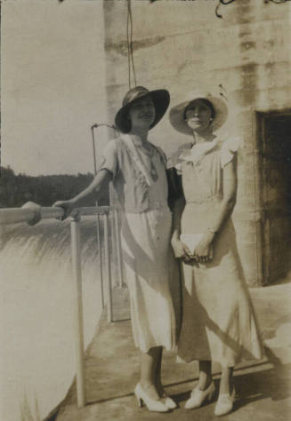 Untitled [Two women in hats stand, one rests hand on rail, one holds purse with both hands]