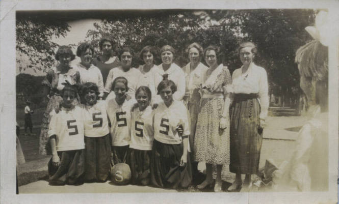 Untitled [Fourteen figures stand together, five people kneel wearing shirts that read 5, two figures on either side appear in the periphery]