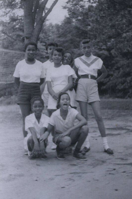 Untitled [Five women stand on beach, two women kneel in the front, volleyball net in background]