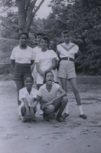 Untitled [Five women stand on beach, two women kneel in the front, volleyball net in background]
