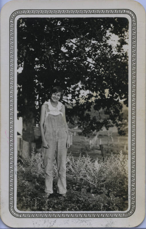 Untitled [Woman stands on field, hands by sides, wears overalls]