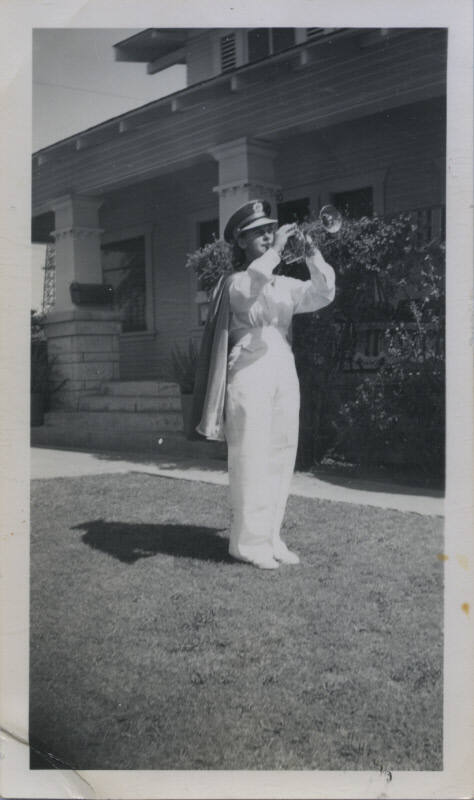 Untitled [Woman in uniform, hat, and cape plays trumpet on house lawn]