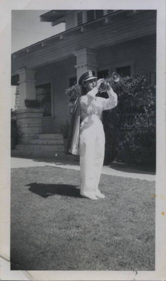 Untitled [Woman in uniform, hat, and cape plays trumpet on house lawn]