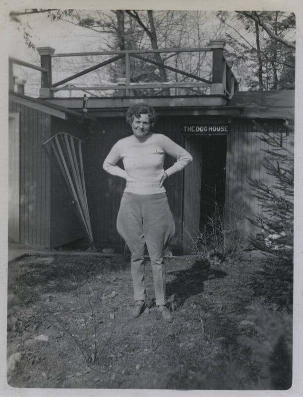 Untitled [Woman stands, hands on hips, house that reads THE DOG HOUSE in background]
