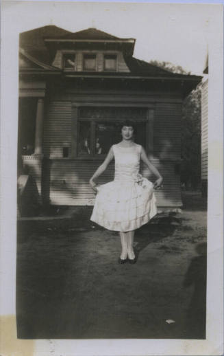 Untitled [Woman stands in front of house, holds up skirt by sides]