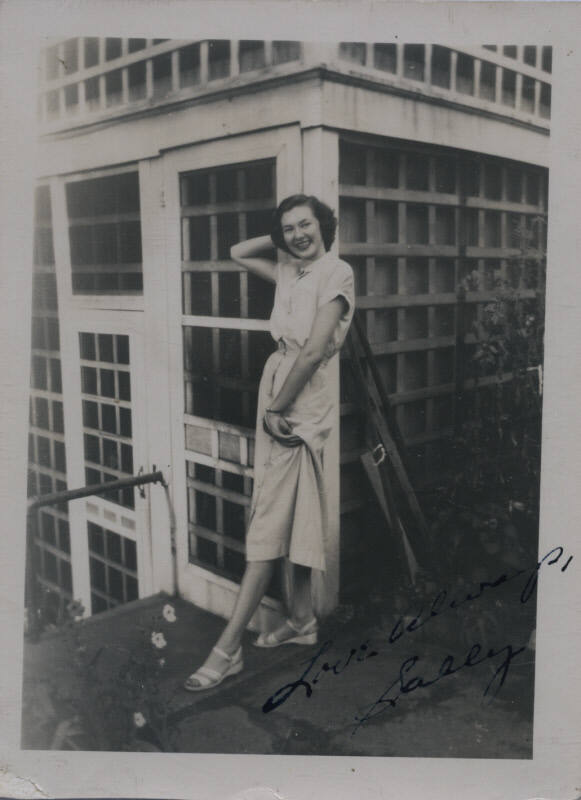 Untitled [Woman stands in terrace, holds dress in one hand and leans back unto building]
