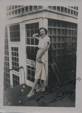 Untitled [Woman stands in terrace, holds dress in one hand and leans back unto building]